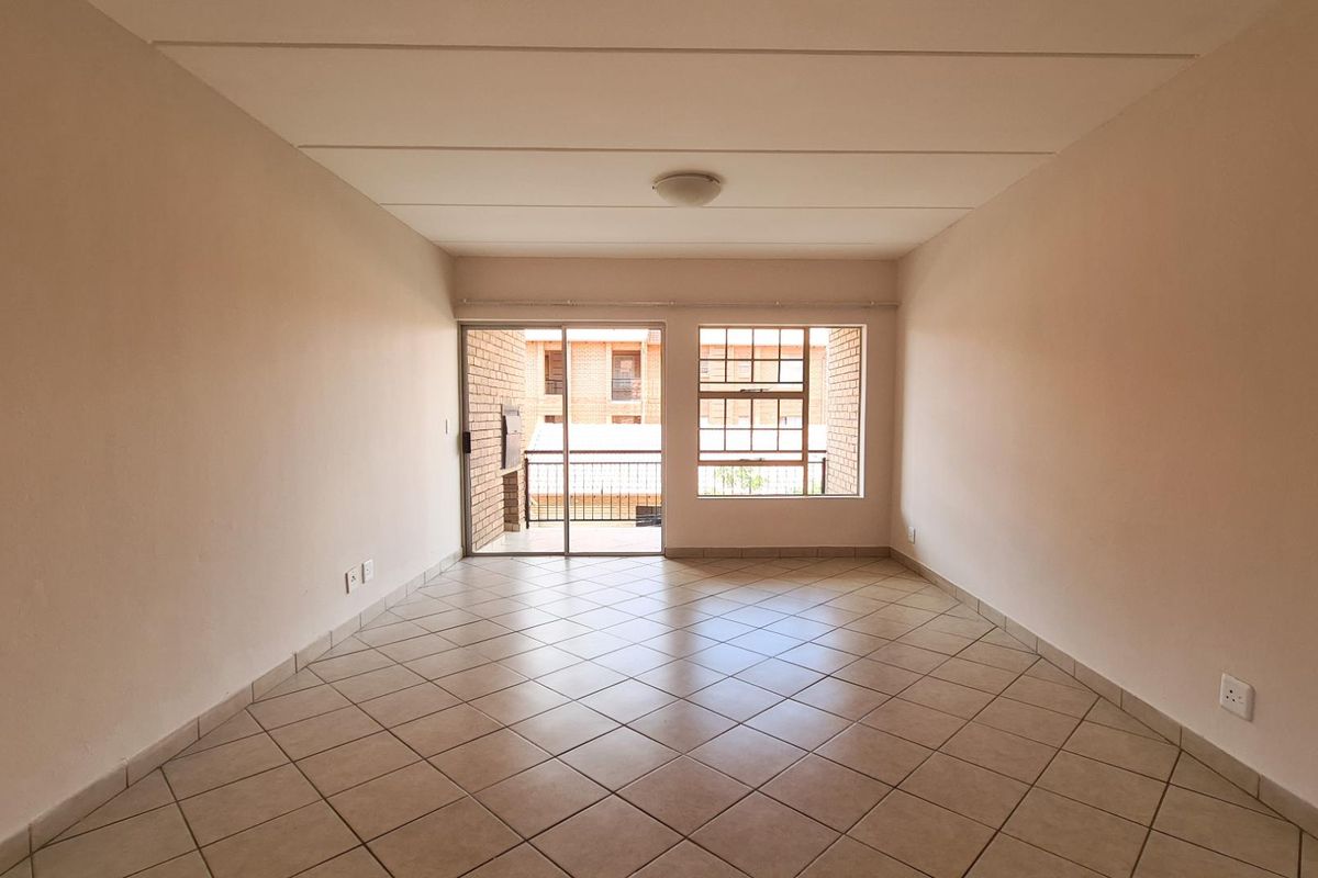 A bright, empty living room featuring neutral tiled flooring, a sliding glass door leading to a balcony, and a large window providing natural light.
