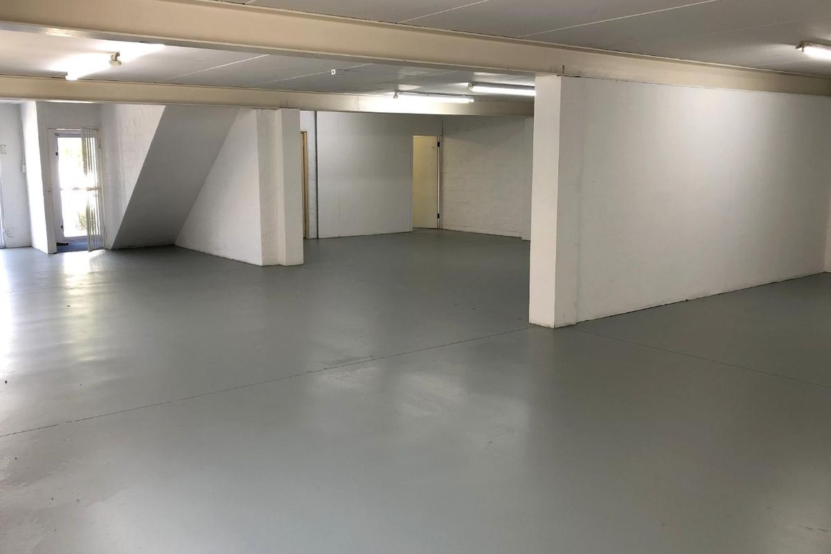 A spacious, open-plan utility or storage area featuring painted concrete flooring, exposed structural beams, and functional overhead lighting.