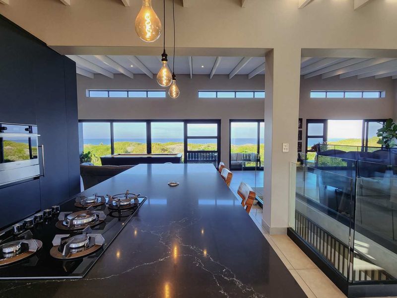 Modern open-plan kitchen and living area featuring a large island with gas hob, sleek cabinetry, exposed beam ceilings, and expansive ocean views.