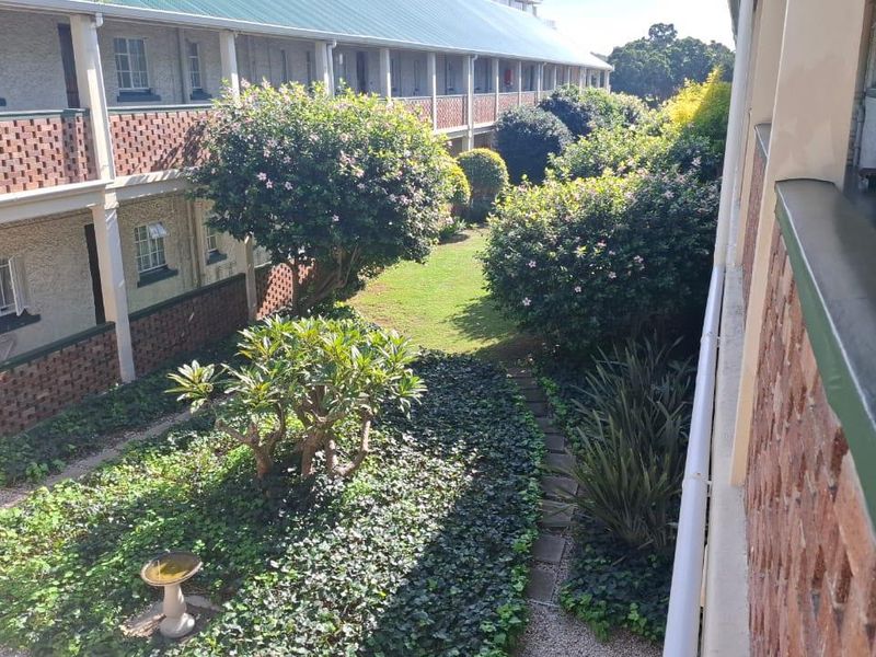 A view of the communal garden area and landscaped grounds situated between apartment building wings, featuring established greenery and paved pathways.