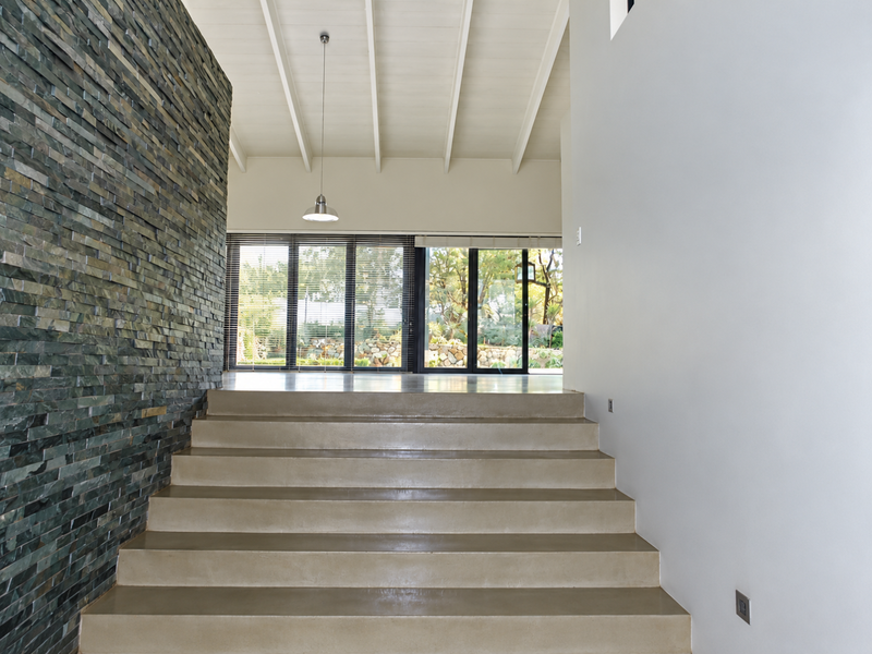 Spacious entrance hall featuring a striking stacked stone feature wall, polished concrete staircase, and high exposed beam ceilings leading to a bright living area.
