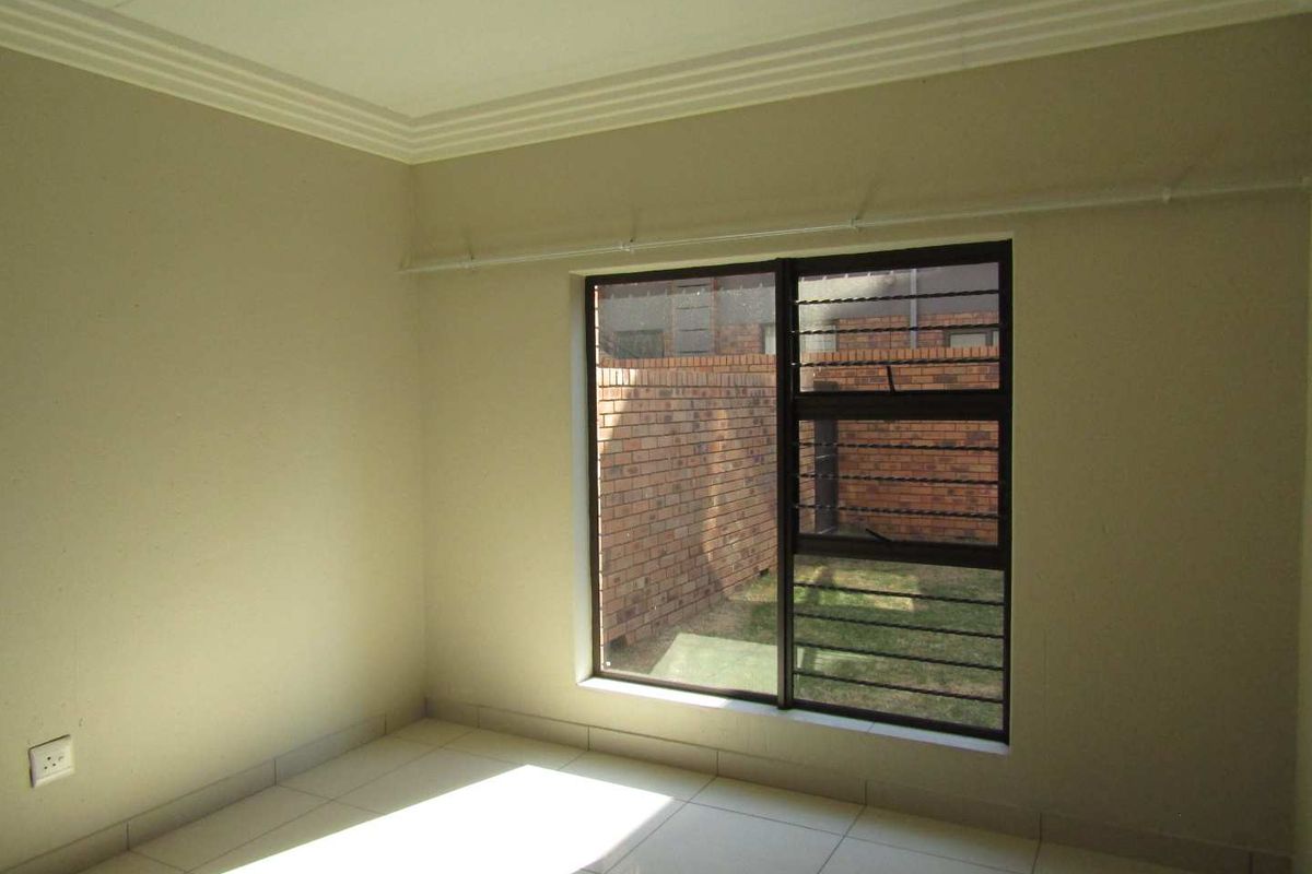A clean, empty bedroom featuring neutral tiled flooring, cornice detailing, and an aluminium window with security bars overlooking a private rear courtyard.