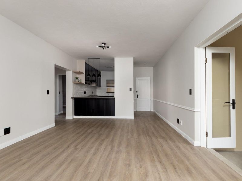 A bright, modern open-plan living area featuring light wood-look laminate flooring, wainscoting, and a sleek, contemporary kitchen with dark cabinetry and subway tile backsplash.
