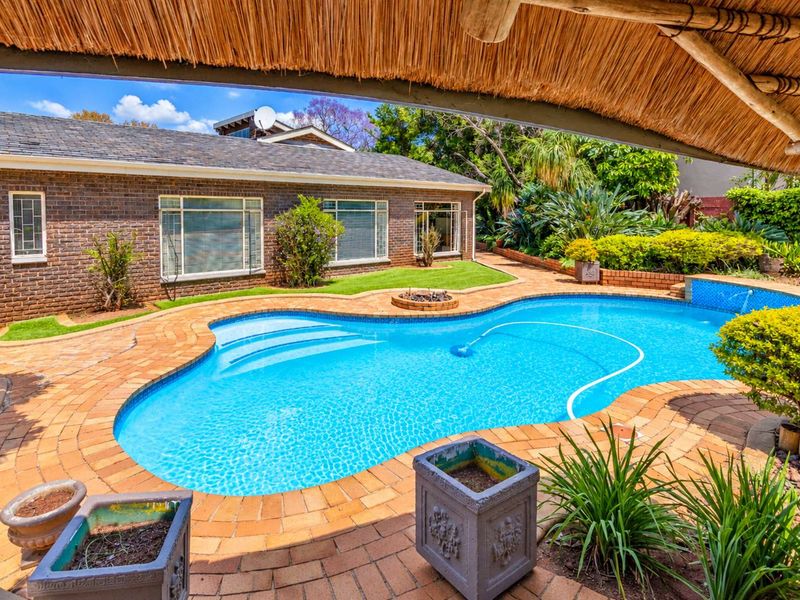 A well-maintained outdoor pool area featuring a swimming pool, brick paving, and a covered thatched patio, set against a landscaped garden.