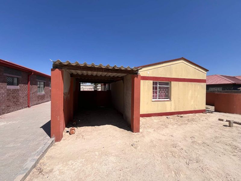 Front view of a house featuring a covered carport, security bars on windows, and a perimeter wall, set on an unpaved lot.