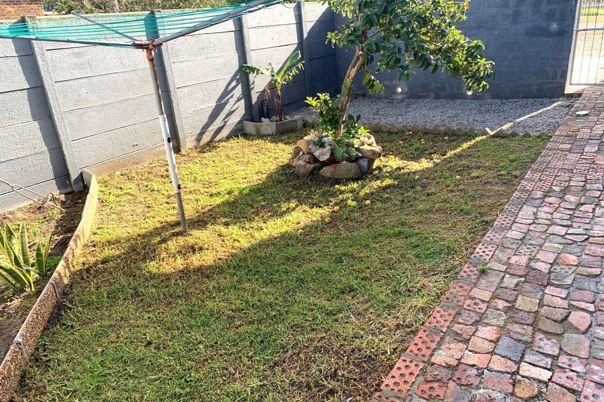 A rear garden area featuring a brick paved pathway, precast concrete boundary walls, and a small lawn area with established trees.
