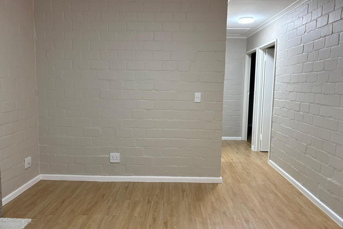 A clean, empty interior space featuring painted brick walls, light wood-look laminate flooring, and access to a hallway.