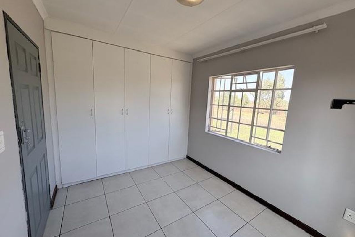 A clean, empty bedroom featuring neutral tiled flooring, built-in storage cupboards, and a steel-framed window providing natural light.