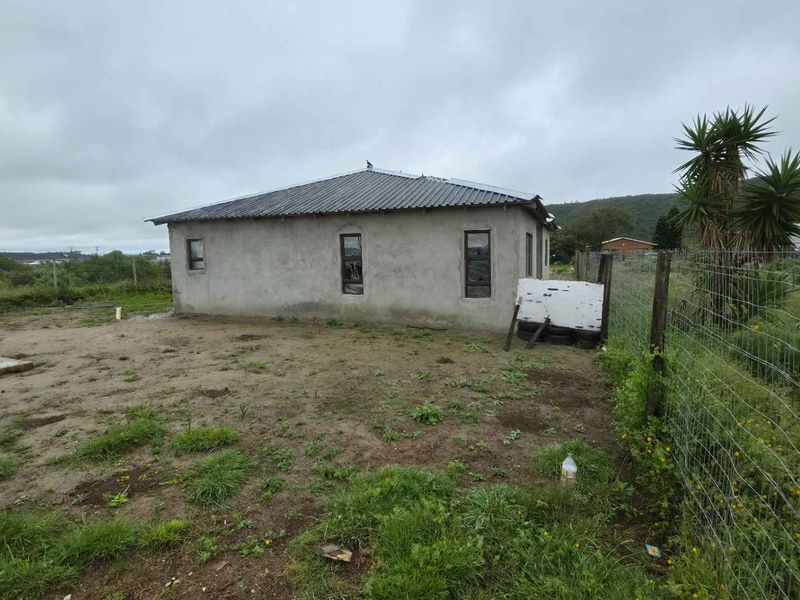 Rear view of a house featuring a corrugated metal roof and unfinished cement plaster exterior walls, set on a large, unlandscaped plot.