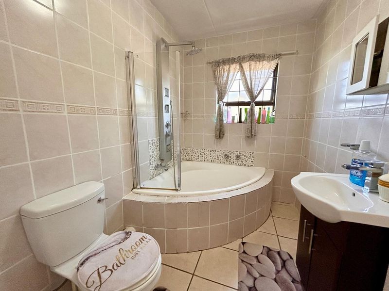 A functional bathroom featuring a corner bathtub with an integrated shower panel, tiled walls and flooring, and a vanity unit with a washbasin.