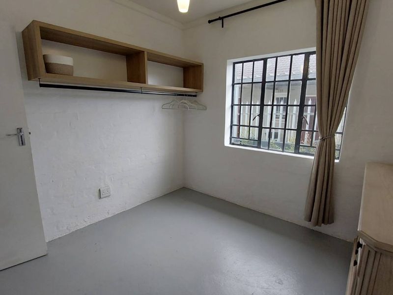 A compact, neutral bedroom featuring painted concrete flooring, a steel-framed window, and built-in wall shelving, offering a clean and functional space.