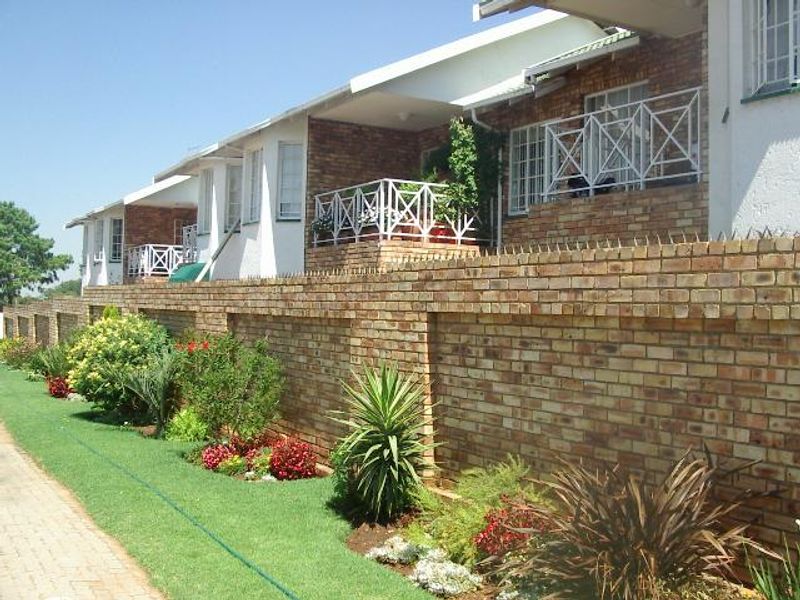 A view of the townhouse complex exterior featuring a brick boundary wall with security spikes, landscaped garden beds, and upper-level balconies with white railings.