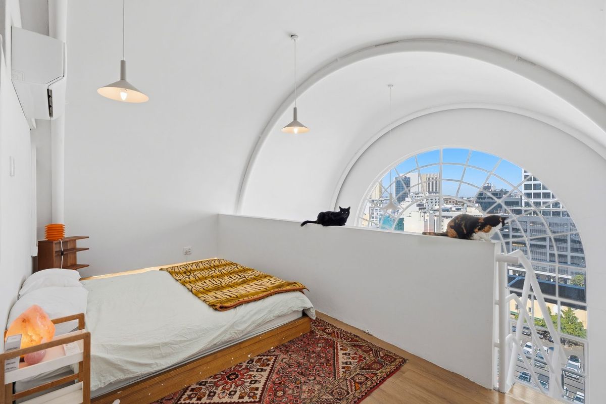 A unique loft-style bedroom featuring a striking arched window with city views, high ceilings, and modern pendant lighting.