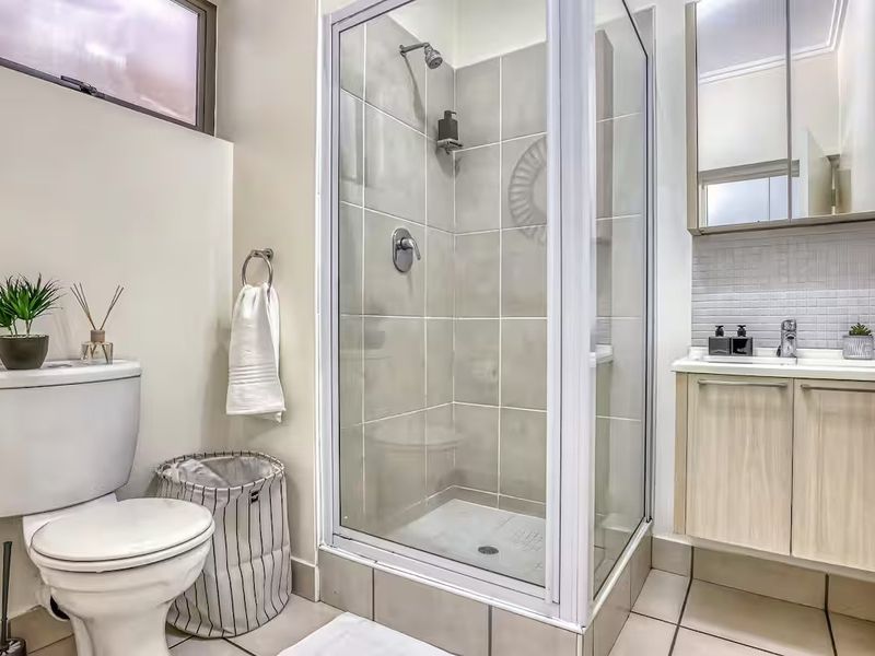 A clean and functional bathroom featuring a glass-enclosed shower, wall-mounted vanity with basin, and a standard toilet with neutral tiled finishes.