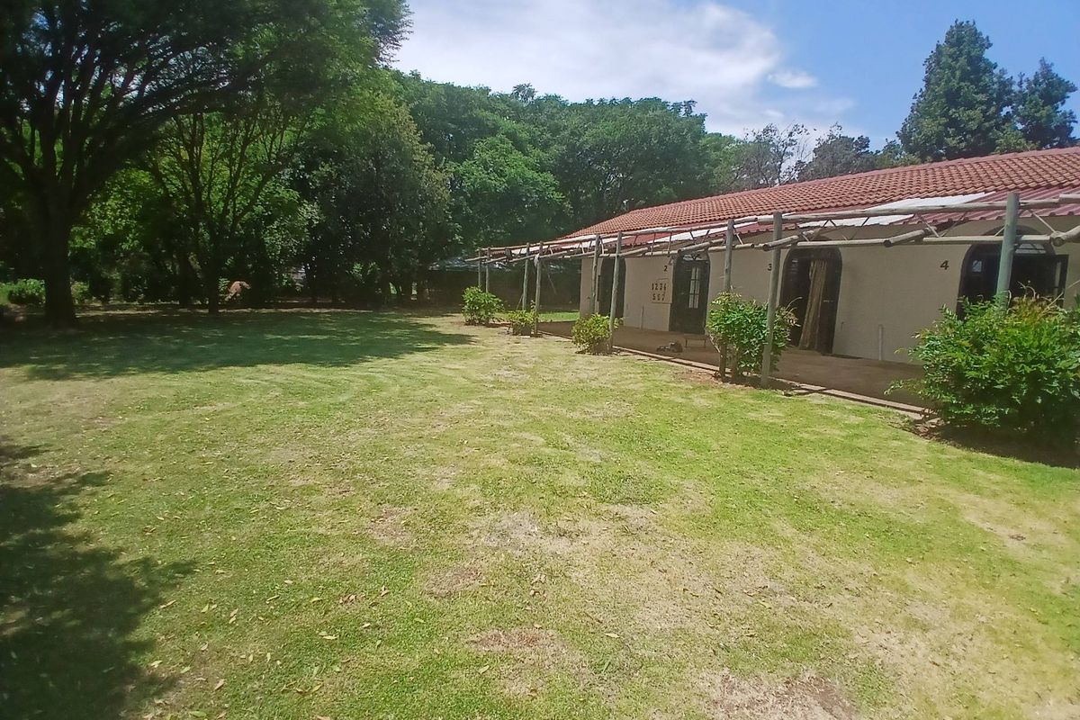 A rear view of the cottage featuring a paved patio area covered by a timber pergola, overlooking a spacious garden with mature trees.