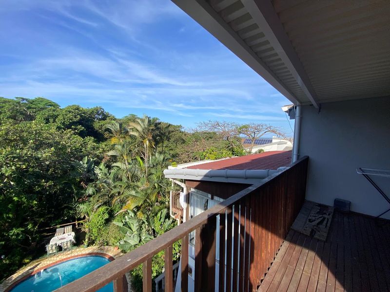 A covered balcony featuring timber decking and a wooden balustrade, offering an elevated view over the surrounding garden and swimming pool area.