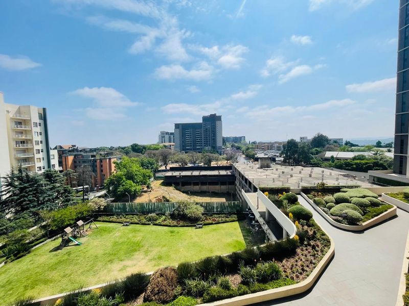 An elevated view overlooking a well-maintained communal garden area with manicured lawns, landscaped shrubbery, and paved pathways within an apartment complex.