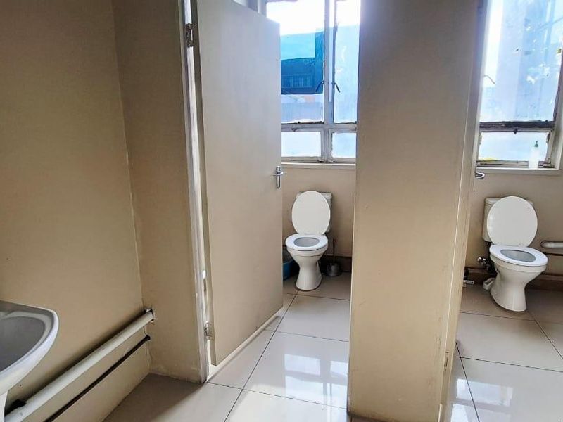 A functional commercial bathroom facility featuring multiple toilet cubicles, pedestal wash basins, and tiled flooring with large windows for natural light.