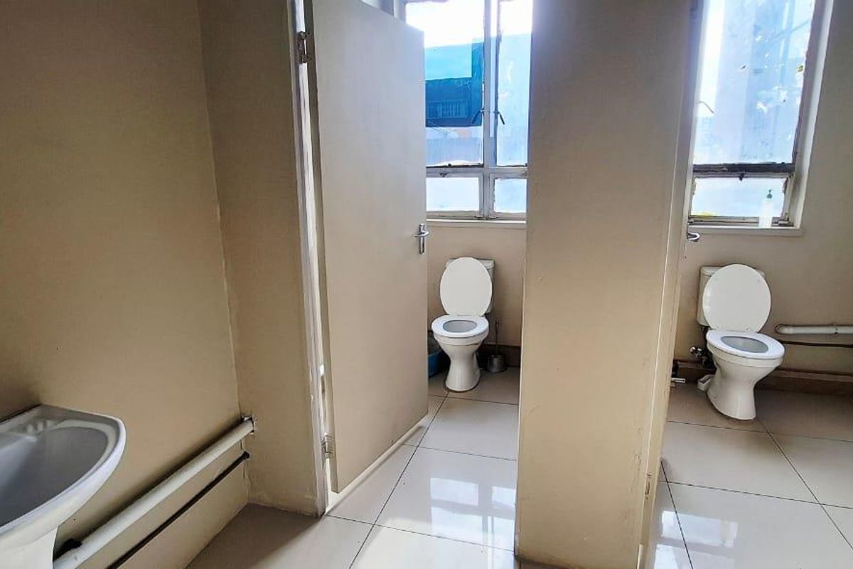 A functional commercial bathroom facility featuring multiple toilet cubicles, pedestal wash basins, and tiled flooring with large windows for natural light.