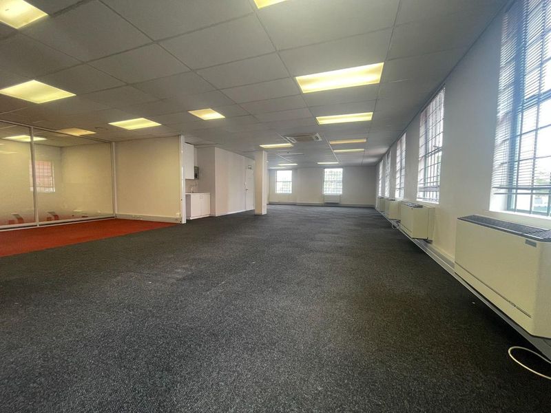 Spacious open-plan office area featuring suspended ceilings, recessed lighting, carpeted flooring, a small kitchenette, and glass-partitioned meeting space.