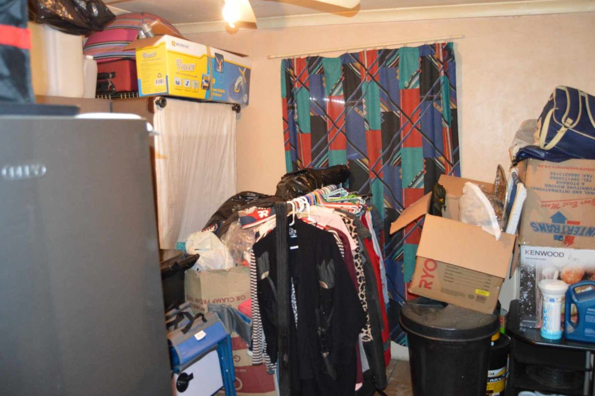 A cluttered utility space or bedroom area featuring a ceiling fan and window with curtains, currently used for storage.
