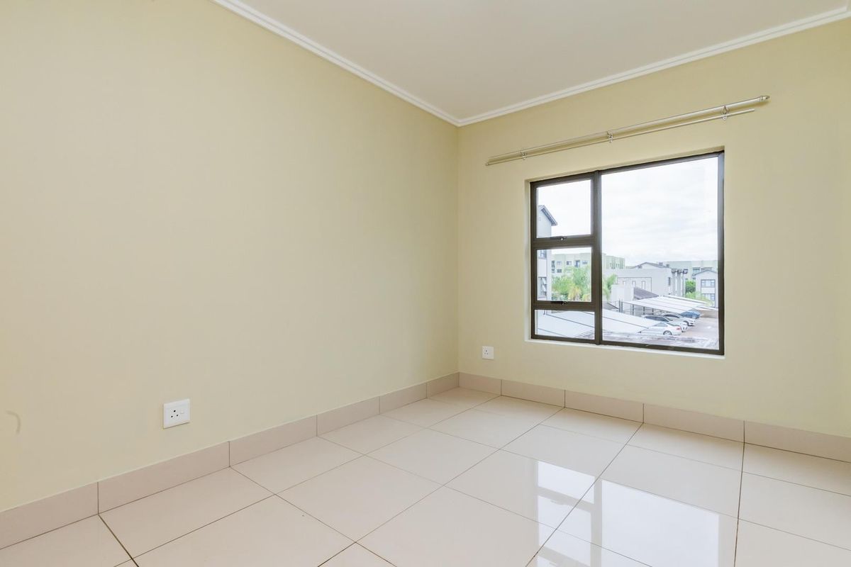 A bright, empty bedroom featuring neutral wall paint, large tiled flooring, and an aluminium-framed window with a view of the complex.