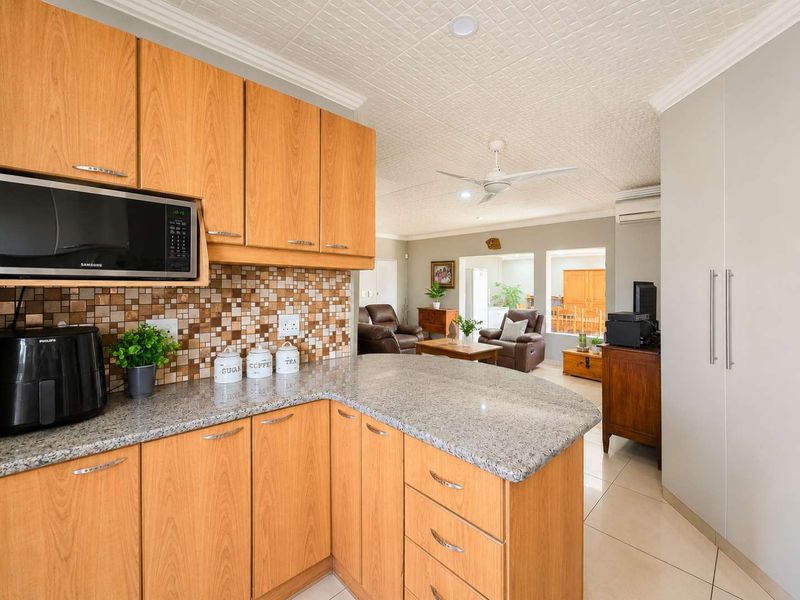 Open-plan kitchen featuring wood-finish cabinetry, granite countertops, and a mosaic backsplash, flowing into a spacious living area with tiled floors and ceiling fan.