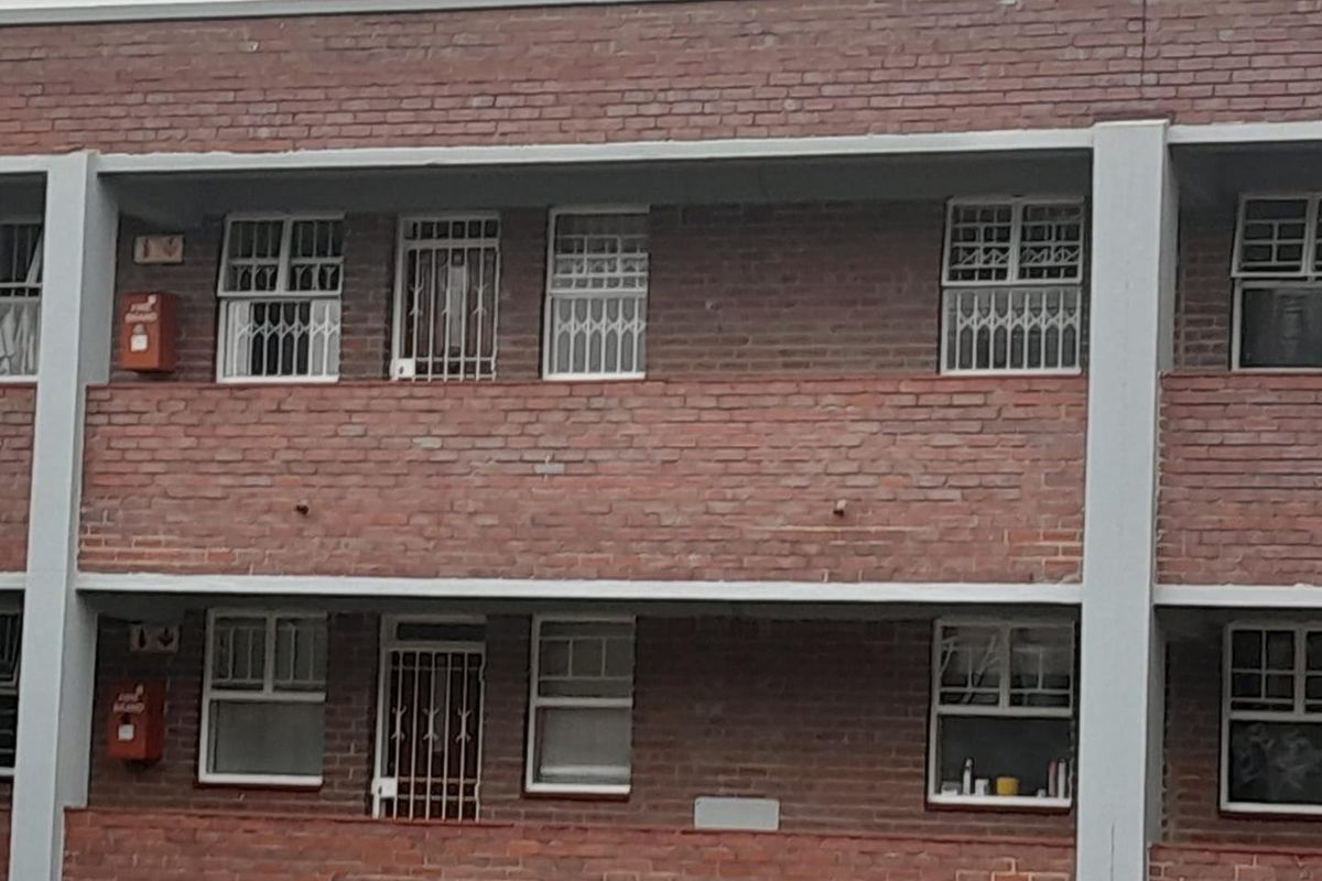 Exterior view of a multi-story apartment building featuring red brick facade, concrete structural elements, and security bars on windows.