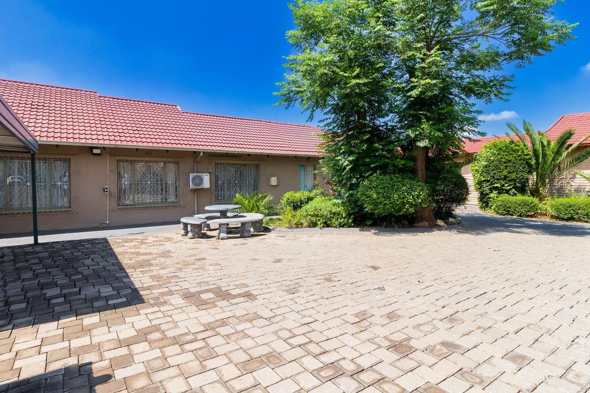 A paved courtyard area featuring a single-story building with a red tiled roof, security window grilles, and established garden landscaping.