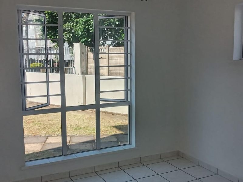 A bright, empty bedroom featuring tiled flooring and large windows with security bars, offering a view of the exterior grounds.