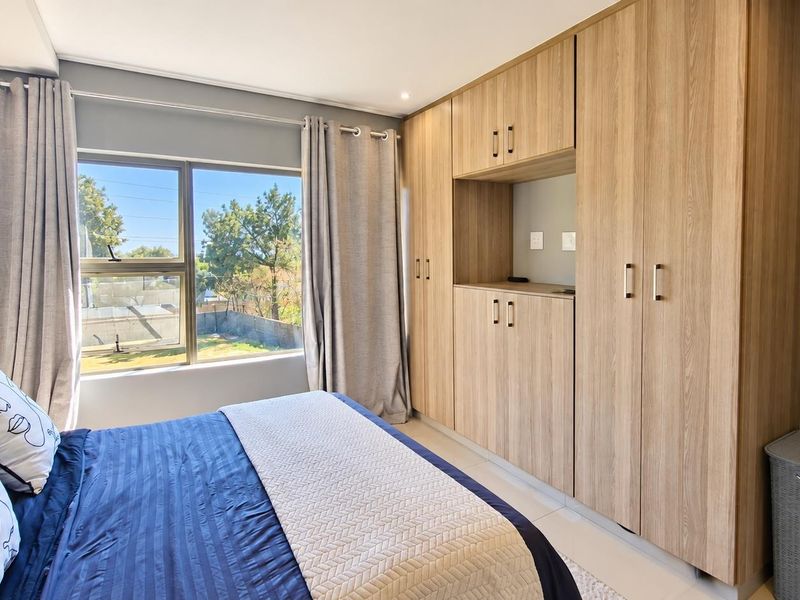 A well-maintained bedroom featuring modern light wood built-in cupboards, tiled flooring, and a large window providing natural light and garden views.
