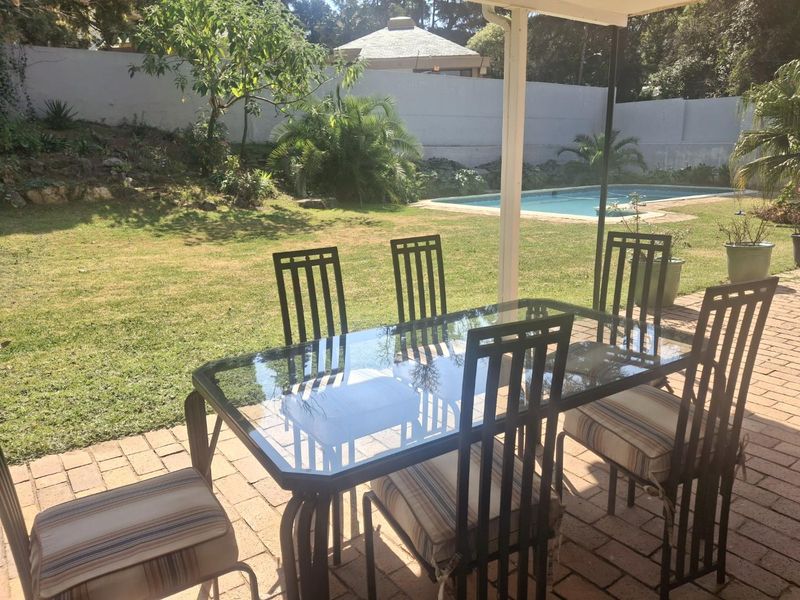 A covered outdoor patio area featuring brick paving, overlooking a private garden and swimming pool.