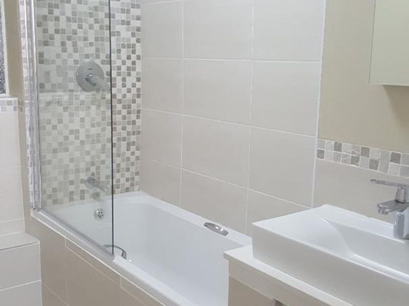 A clean, modern bathroom featuring a bathtub with a glass shower screen, mosaic tile accents, and a sleek vanity basin.