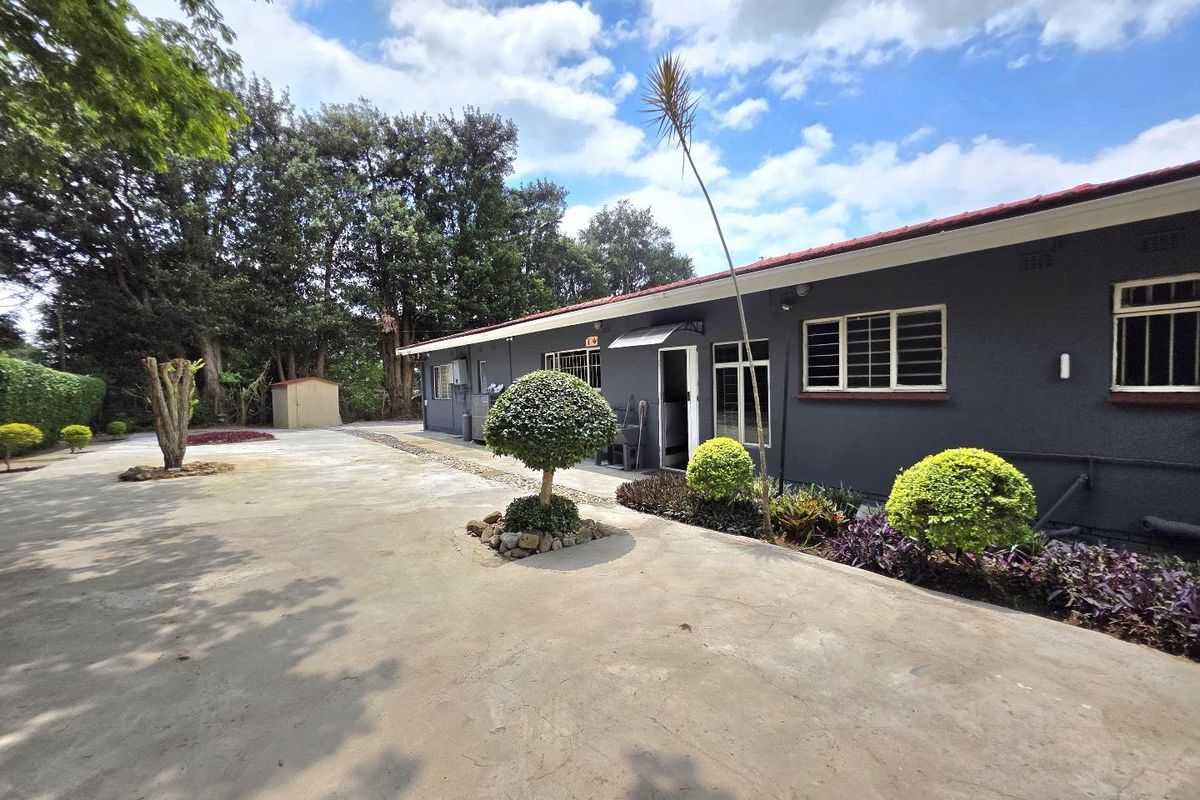 A view of the rear exterior of the house featuring a large concrete driveway, landscaped garden beds, and mature trees.