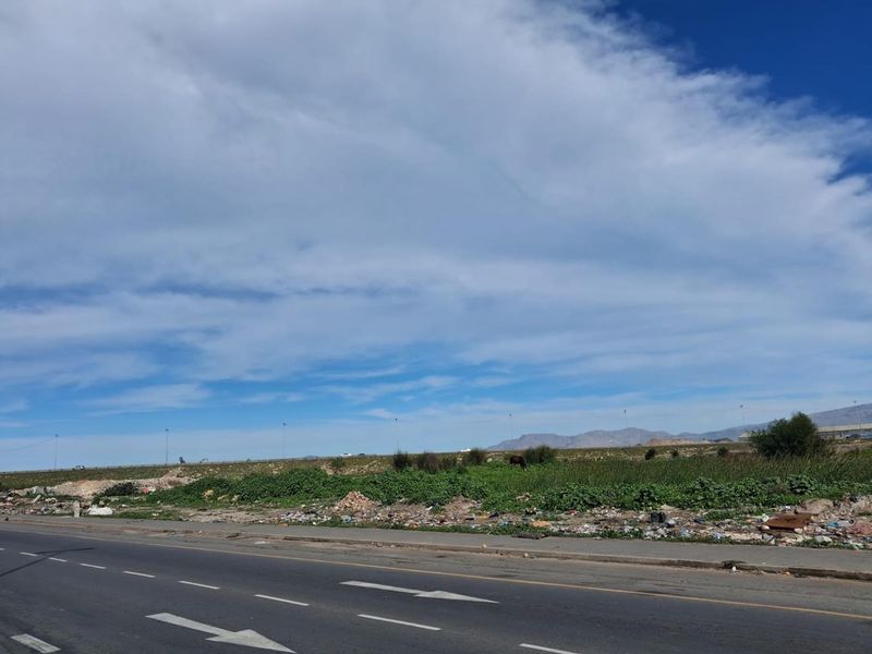 A street-level view of an undeveloped agricultural land parcel adjacent to a paved road, showing natural vegetation and surrounding landscape.