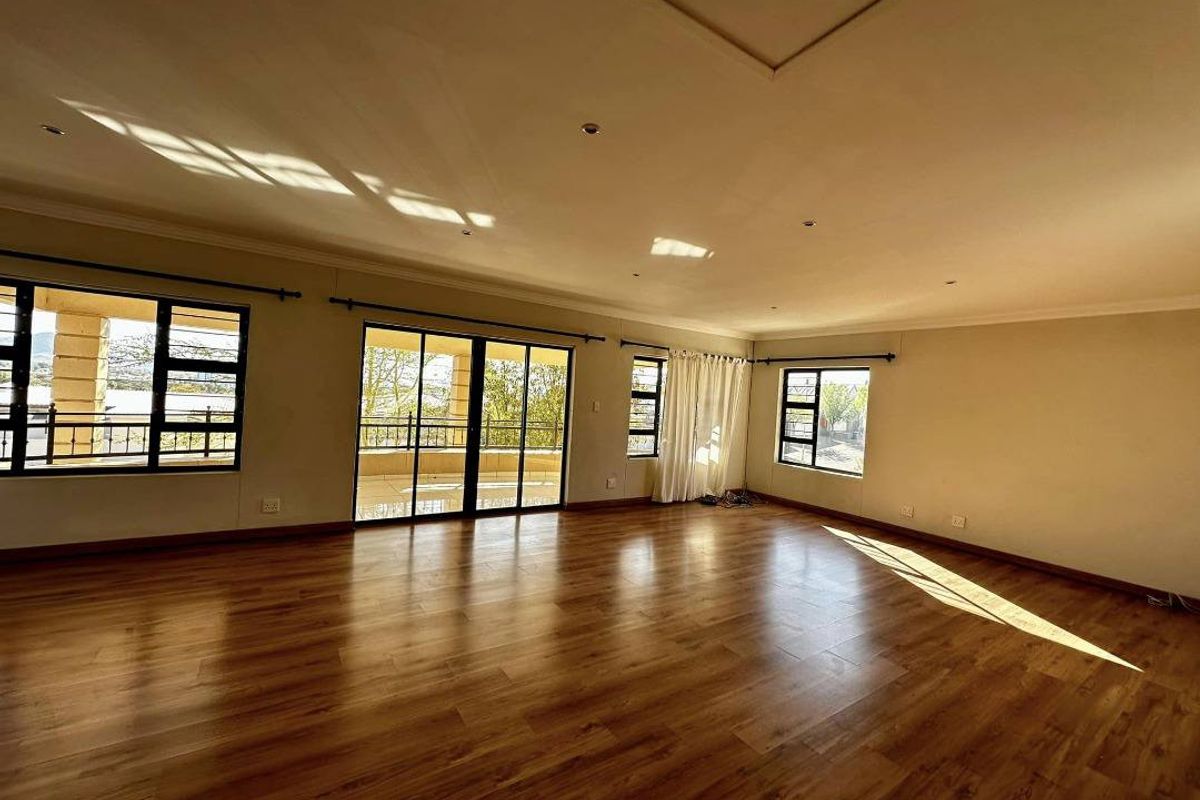 A spacious, sunlit living area featuring warm-toned laminate flooring, large windows, and sliding glass doors that open onto an outdoor balcony.