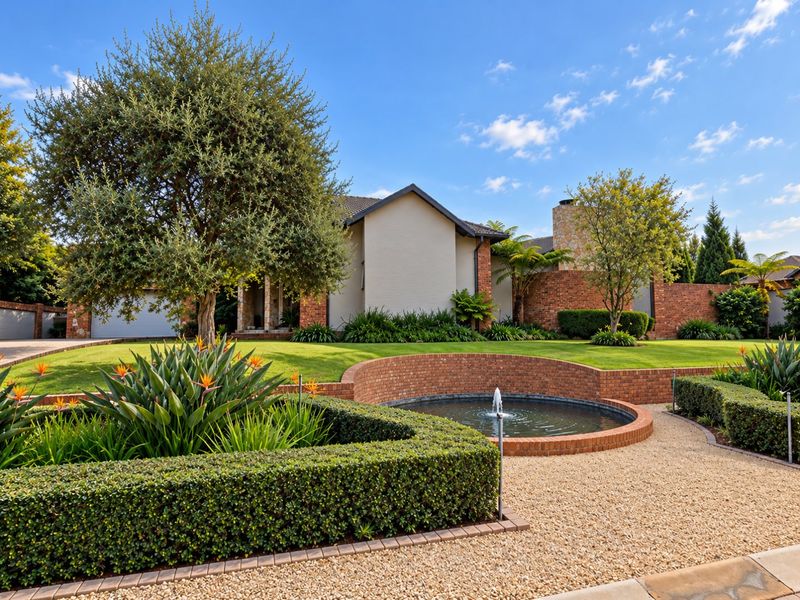 A beautifully landscaped front garden featuring a circular brick fountain, manicured hedges, and a well-maintained brick and stone house facade.