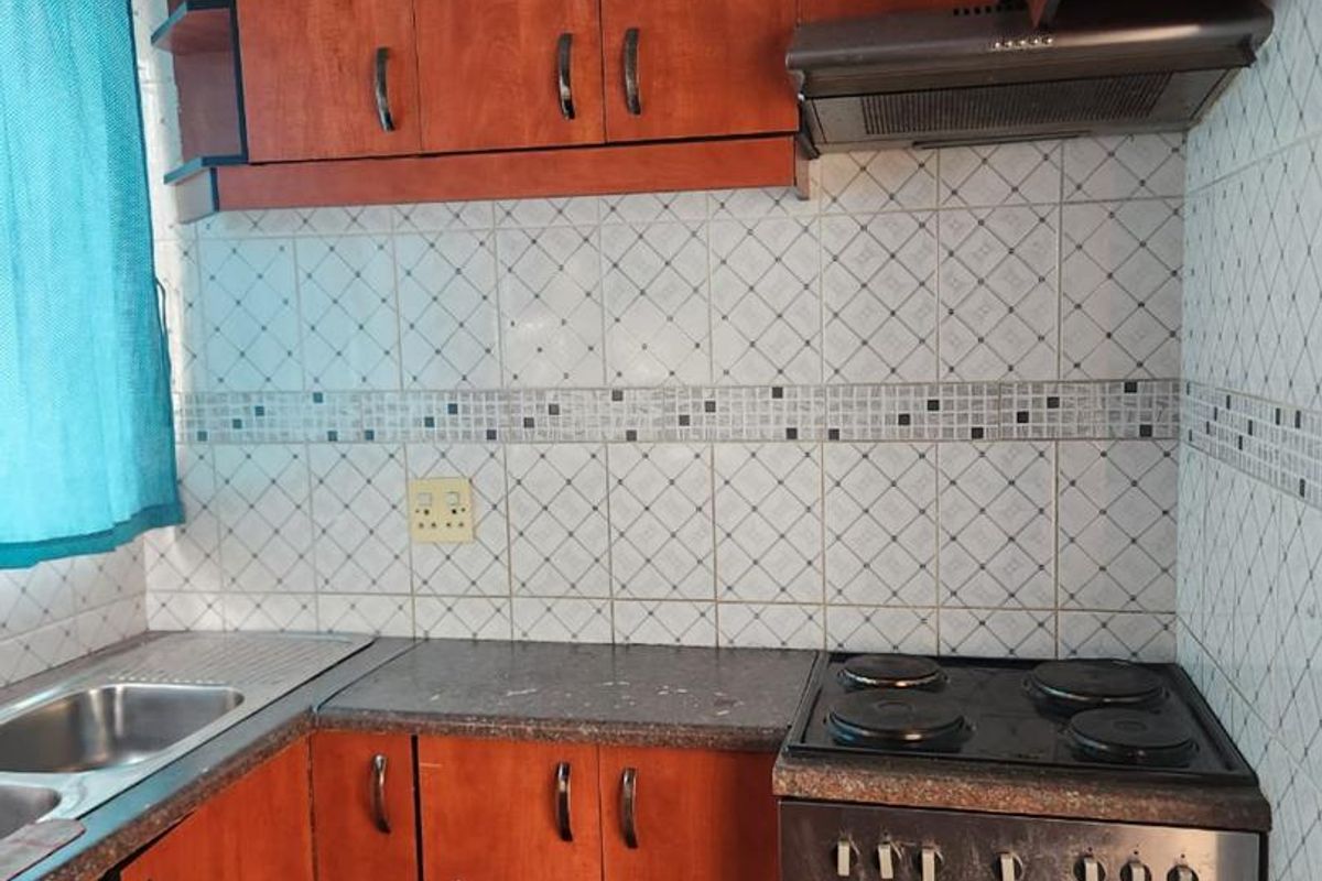 A functional kitchen featuring wooden cabinetry, a built-in oven with hob, extractor fan, and a stainless steel sink with tiled backsplash.