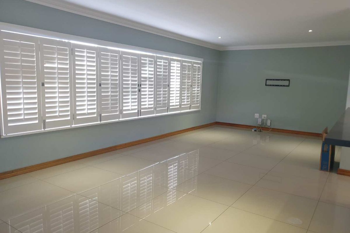 Spacious, empty living area featuring large white plantation shutters, polished porcelain floor tiles, and neutral wall tones with recessed lighting.