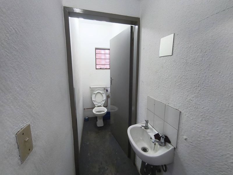 A functional industrial-style bathroom featuring a wall-mounted basin with tiled splashback and a separate toilet area with a small louvered window.