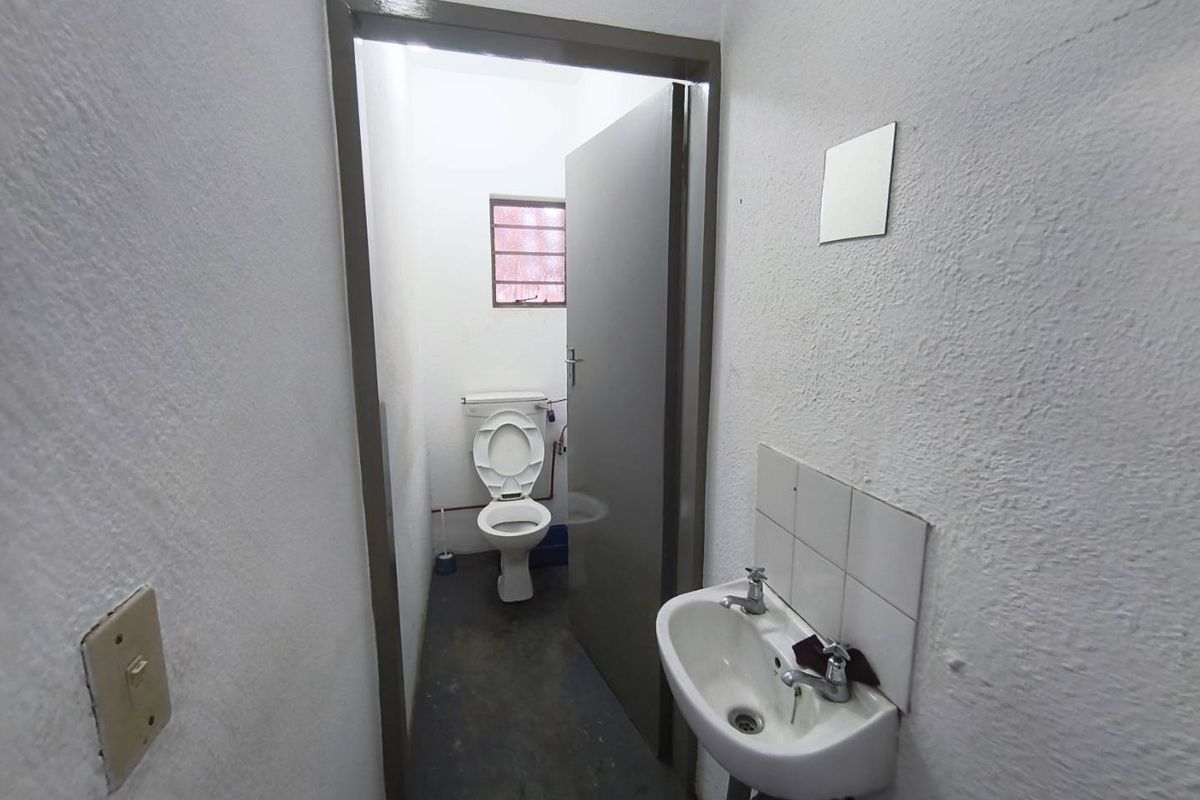 A functional industrial-style bathroom featuring a wall-mounted basin with tiled splashback and a separate toilet area with a small louvered window.