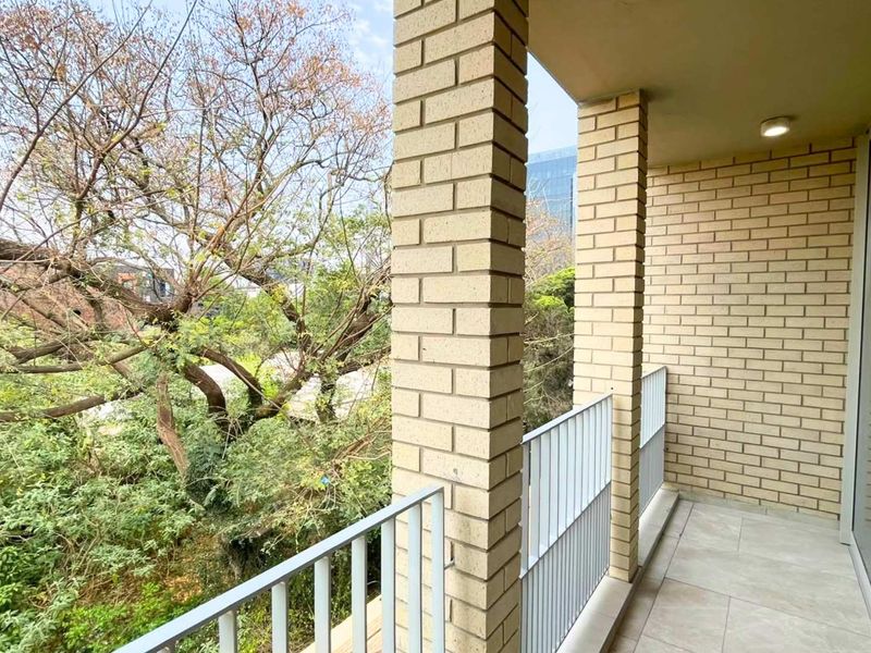 A clean, covered balcony featuring brick columns, metal railings, and tiled flooring, offering a pleasant view of surrounding trees.