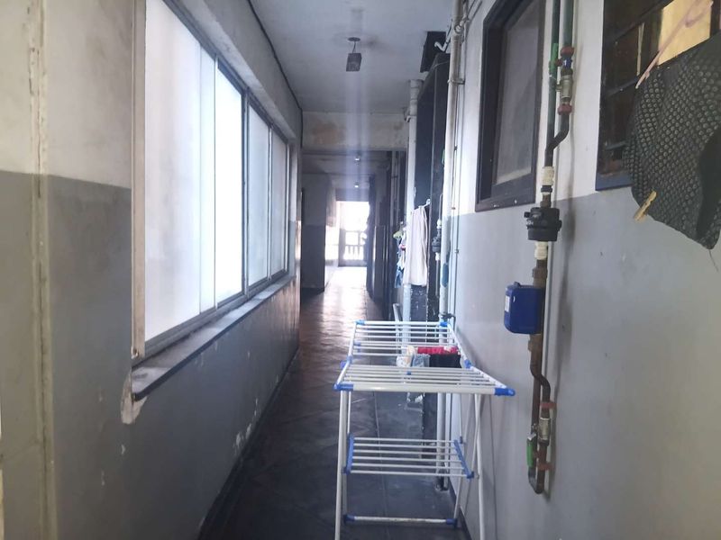 A narrow apartment hallway featuring tiled flooring, large windows, and exposed utility piping, showing signs of wear and requiring cosmetic updates.