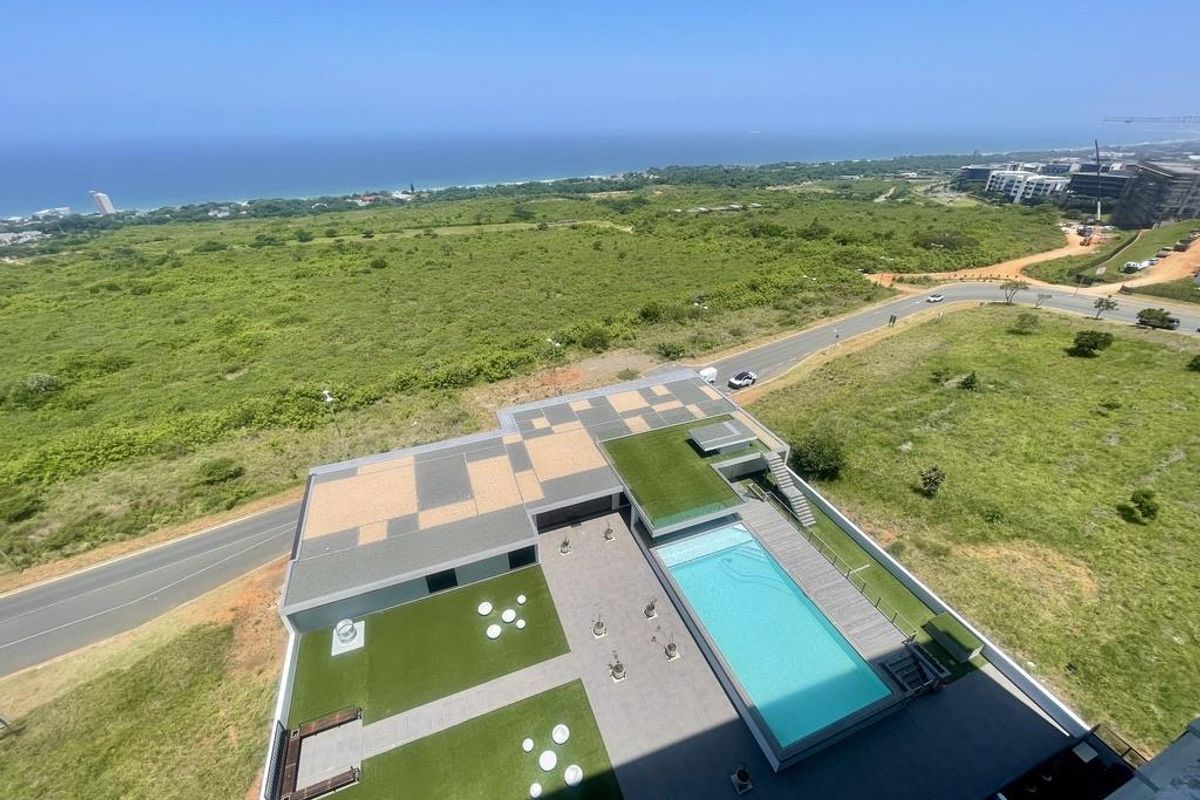 Aerial view of a modern apartment complex featuring a swimming pool, landscaped garden areas, and paved outdoor recreational space with coastal views.