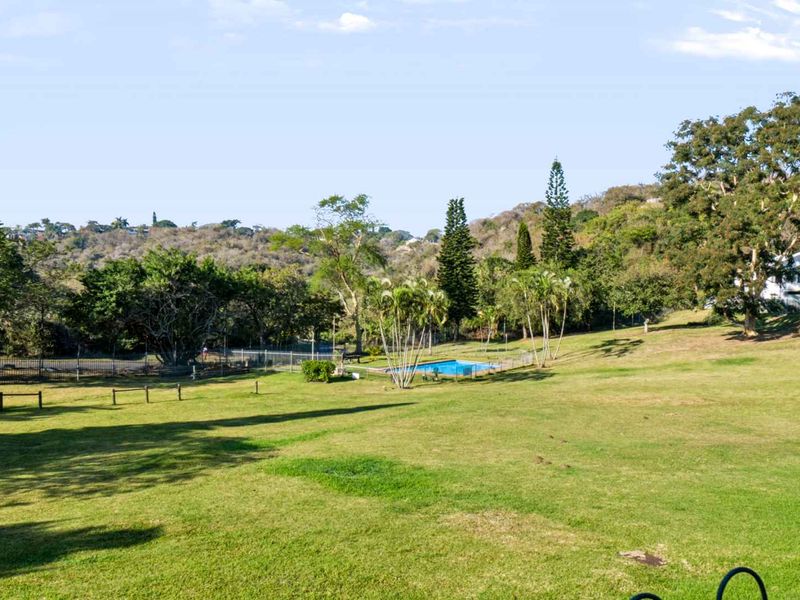 A scenic view of the expansive communal garden area featuring a swimming pool and mature trees set against a hilly landscape.