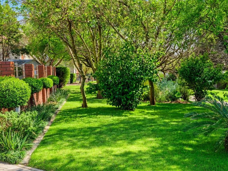 A well-maintained garden featuring a brick boundary wall, mature trees, lush lawn, and a paved driveway with an automated gate.