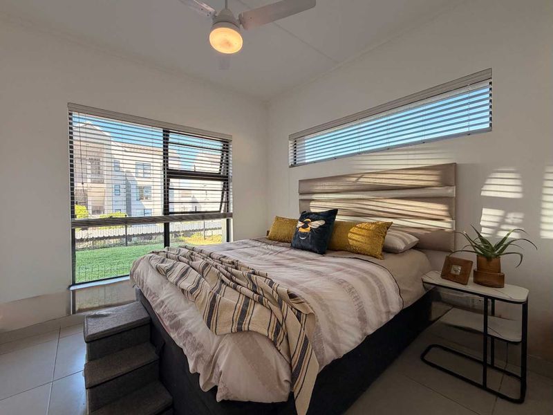 A bright and well-maintained bedroom featuring tiled flooring, a ceiling fan, and dual-aspect windows with fitted horizontal blinds.