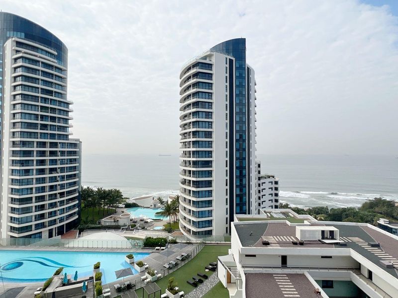 Elevated view of modern high-rise apartment towers overlooking a coastal swimming pool area and the ocean.