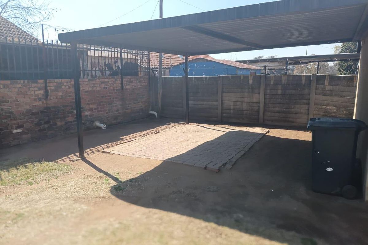 A rear outdoor area featuring a metal carport structure, paved section, and perimeter boundary walls with security fencing.