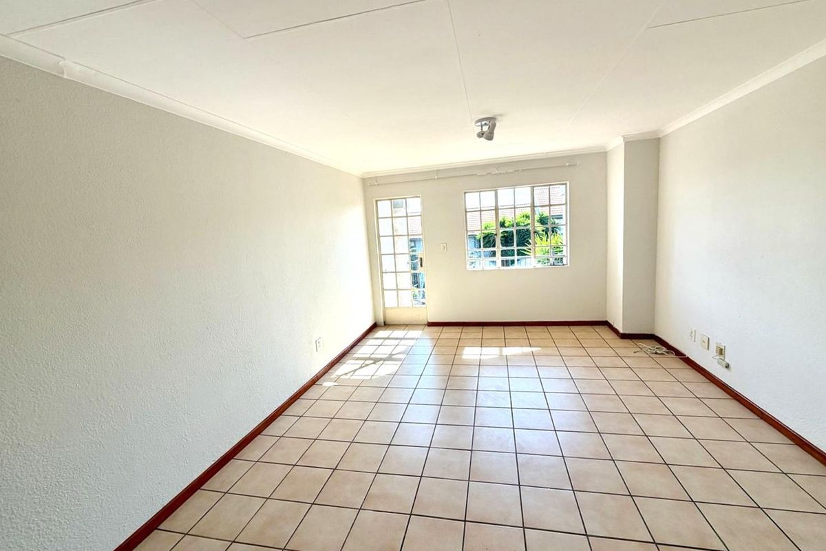 A bright, empty living area featuring neutral tiled flooring, large windows, and a glass-paneled door providing natural light and outdoor access.